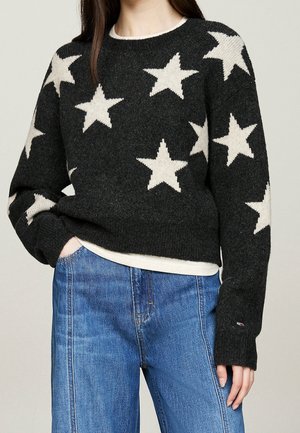 Person iført en sort sweater med store hvide stjerner kombineret med blå jeans, stående med den ene hånd i lommen mod en neutral baggrund.