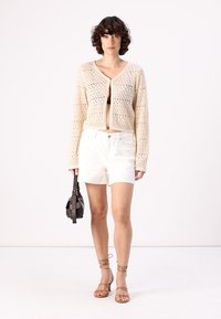 Κροπ πλεκτό καζάκ με V λαιμόκοψη, συνδυασμένο με λευκά σορτς. Το μοντέλο κρατά μια μικρή μαύρη τσάντα και φοράει καφέ λουράκια σανδάλια.