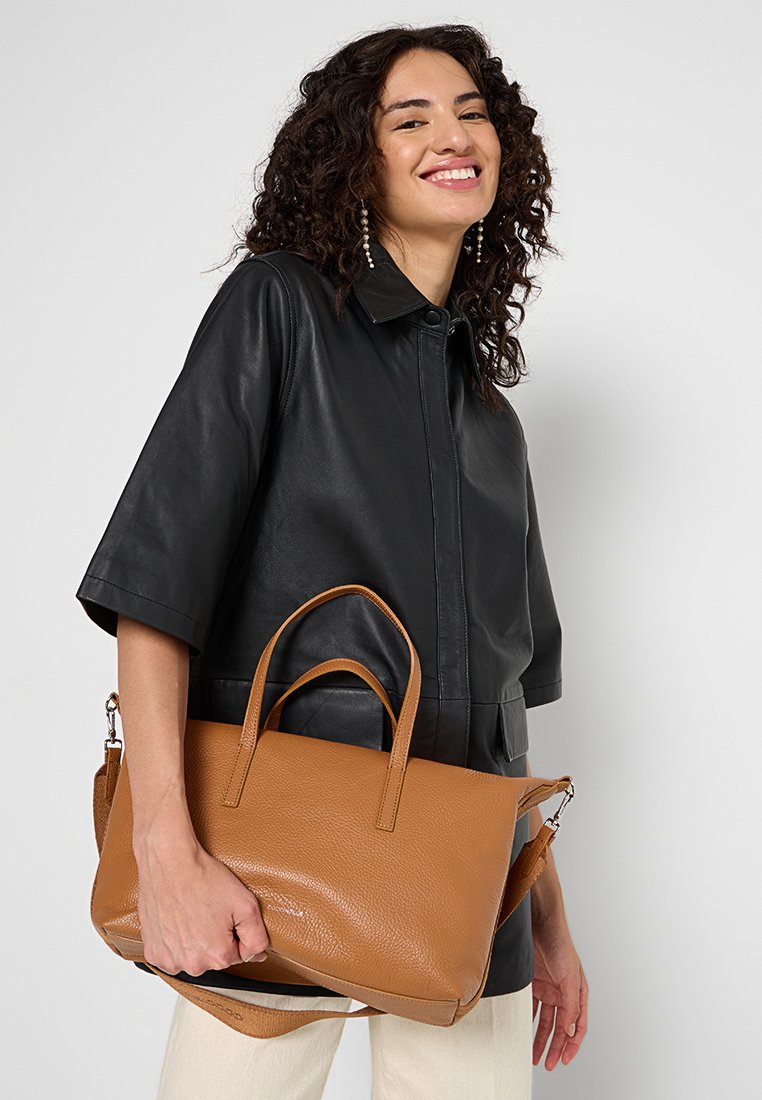 Brązowa skórzana torba tote z podwójnymi uchwytami, odpinanym paskiem na ramię i delikatną fakturą. Tło jest jednolicie białe.