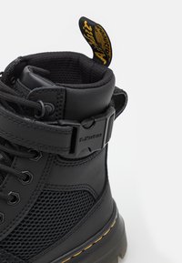 Botte montante noire avec un mélange de cuir lisse et de panneaux en mesh, présentant une pointe arrondie, un laçage, une sangle ajustable et des accents jaunes.