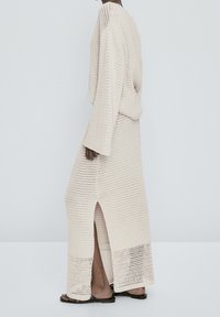 Personne portant une jupe longue en maille crochetée couleur crème et un pull oversize assorti avec une fente latérale, debout devant un fond gris clair.