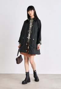 Dark green waxed jacket with gold hardware, brown collar, two patch pockets, layered over a plaid shirt and brown skirt; paired with black boots.