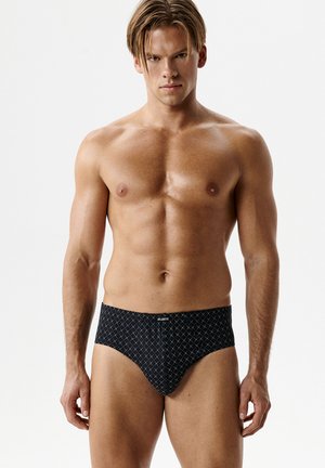 Mężczyzna bez koszulki o jasnobrązowych włosach, ubrany w czarne bokserki z białym wzorem w kratę, stojący na tle jednolitego białego tła.