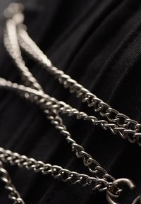 Silberne Metallkette mit ineinander verschlungenen Gliedern, die in Größe und Form variieren, auf einer schwarzen Stoffoberfläche liegend. Enthält ein Verschlussdetail.