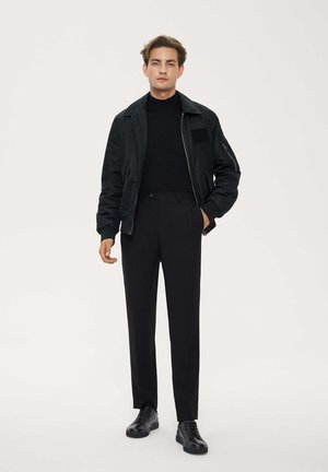 Chaqueta bomber negra con cuello alzado, combinada con un suéter de cuello alto negro y pantalones de vestir. Tejido texturizado y diseño minimalista. Zapatos negros.