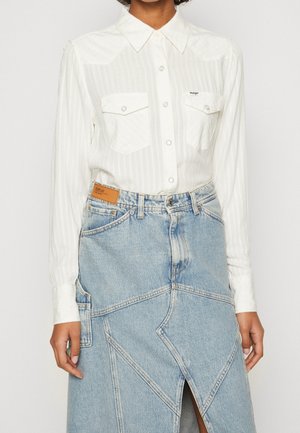 Weißes langärmliges Hemd mit Knöpfen und zwei Brusttaschen, kombiniert mit einem hoch taillierten, hellblauen Jeansrock mit geometrischen Nähten.