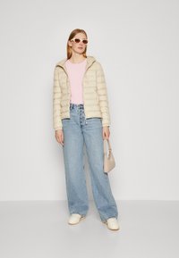 Beige polstret jakke med hette, rosa genser, lyseblå vide bukser og hvite joggesko. Holder en liten beige håndveske.
