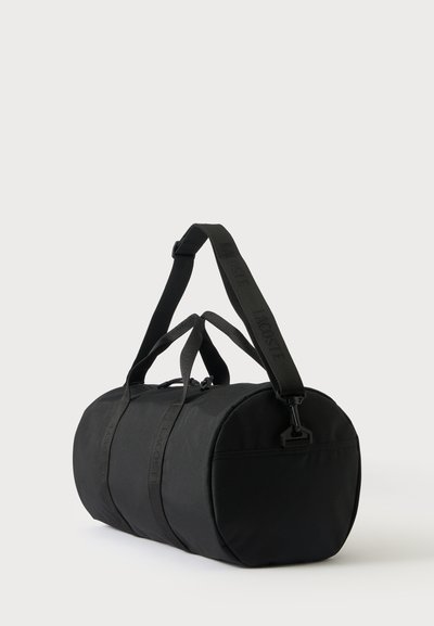Sac de sport cylindrique noir avec de courtes poignées, une bandoulière amovible et une poche zippée à une extrémité, présenté sur un fond uni.