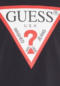Svart T-skjorte med en stor hvit og rød trekantet logo. Teksten inkluderer "GUESS U.S.A," "WASHED," "JEANS," og numrene 1201, 1203.