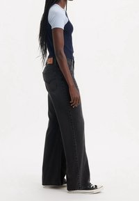 T-paita tummansinisestä ja vaaleansinisestä lyhythihaisesta kankaasta, mustat korkeavyötäröiset leveälahkeiset farkut ja mustat lenkkarit; merkki näkyvissä farkkujen takana.