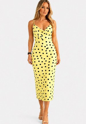 Femme aux longs cheveux blonds portant une robe midi jaune sans manches à pois noirs et des sandales à talons couleur nude, debout devant un fond blanc.