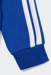 Les poignets du sweat-shirt bleu présentent un tissu texturé avec des bords côtelés et un design à trois rayures blanches sur le côté.