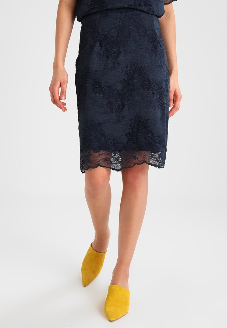 Jupe crayon en dentelle marine avec un ourlet festonné, ornée de motifs floraux. Associée à des chaussures pointues en daim jaune.