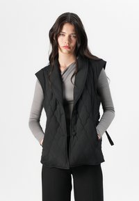 Gilet matelassé noir avec un col, des boutons à l'avant et des liens sur les côtés. Porté par-dessus un haut ajusté à manches longues gris. Texture de tissu lisse.