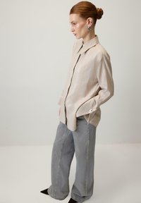 Žena s crvenom kosom u punđu, obučena u bež košulju s prugicama, sive hlače širokih nogavica, srebrne naušnice na obruče, stoji naslonjena na jednostavnu svijetlu pozadinu.