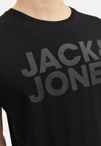 Black cotton t-shirt featuring a large, dark gray "JACK & JONES" logo across the chest. Crew neck and short sleeves. Soft texture.
