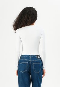 White long-sleeve bodysuit paired with dark blue denim jeans featuring a CK logo on the waistband and two back pockets. Smooth texture.