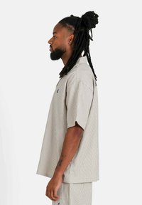 Camisa de manga corta a rayas en beige y gris claro, con un corte relajado y un pequeño detalle de logo. Textura suave, diseño de cuello abierto.