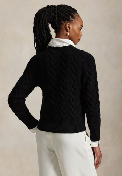 Fonott hajú nő fekete kötött mintás pulóvert visel fehér galléros ing és fehér nadrág fölött, oldalra néz egy semleges háttér előtt.