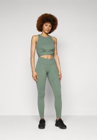 Set sportivo verde composto da un top corto con dettaglio annodato e leggings a vita alta. Entrambi i capi hanno una texture liscia e aderente.