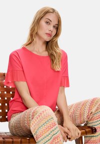Coral top with pleated short sleeves, paired with multicolored patterned pants featuring zigzag design. Model seated on a wooden chair.