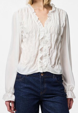 Frau trägt eine weiße Bluse mit langen Ärmeln, Rüschen vorne und an den Manschetten, kombiniert mit dunkelblauer Jeans.