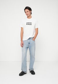 Biała bawełniana koszulka z czarnym napisem "Calvin Klein", zestawiona z jasnoniebieskimi dżinsami. Model nosi czarne trampki, stojąc na jednolitym tle.