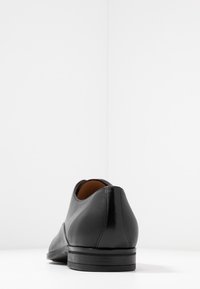 Chaussure habillée en cuir noir avec une finition lisse, un bout arrondi et un profil bas. Présente une texture subtilement côtelée sur le côté et un talon élégant.