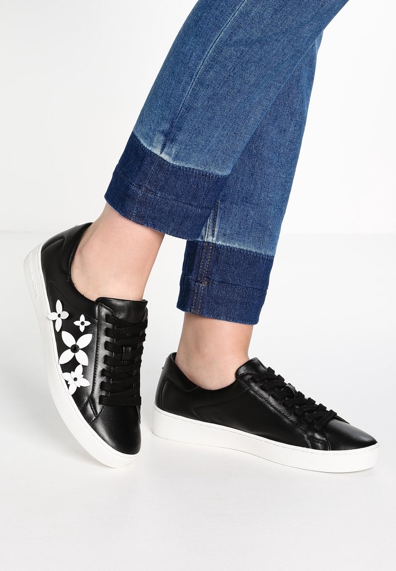 Black leather sneakers with white floral accents, round toe, and rubber sole, paired with dark blue cuffed jeans.