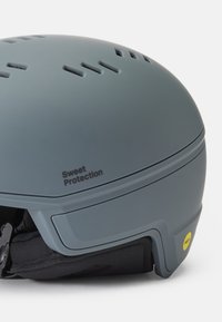 Casco de esquí gris con agujeros de ventilación, acabado mate y pegatina amarilla de MIPS. Presenta una correa para la barbilla acolchada y la marca "Sweet Protection".