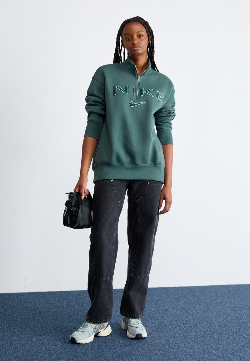 Sweatshirt Nike vert avec demi-fermeture éclair, présentant un logo vert clair. Associé à un jean noir et des baskets bleu clair. Petit sac noir en main.