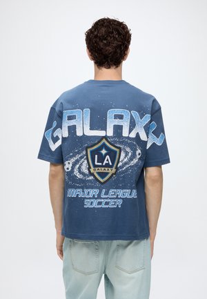 Homme portant un t-shirt bleu LA Galaxy Major League Soccer avec un motif galaxie dans le dos, debout devant un fond blanc.