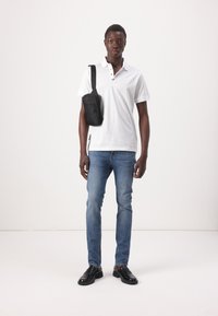 Witte polo met een drieknoopsluiting, gecombineerd met lichtblauwe spijkerbroek en zwarte schoenen. Een kleine zwarte schoudertas hangt over de schouder.