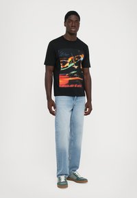 Man wearing black graphic t-shirt, light blue jeans, and green sneakers standing against a plain white background.