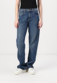 Jean bleu à jambe large en denim, avec une fermeture à bouton, cinq poches et un léger délavage sur le tissu. Associé à des baskets blanches.