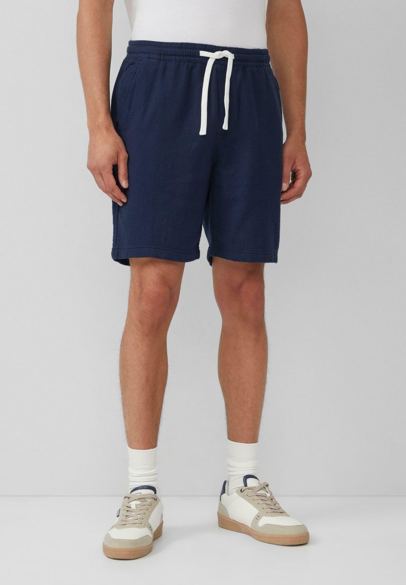 Shorts bleu marine avec une taille élastique et un cordon blanc, présentant un tissu texturé et des poches latérales. Portés avec des chaussettes blanches et des baskets.
