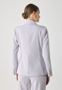Blazer tailored di colore viola chiaro con una silhouette aderente, spalle strutturate e dettagli in bottone dorato sulle maniche. Texture liscia con cuciture minime.