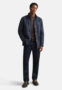 Tmavě modrá prošívaná bunda se zapínáním na patentky, nošená přes hnědý svetr, doplněná tmavými džínami a hnědými koženými botami na šněrování. Bez viditelných vzorů.