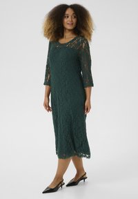 Green lace midi dress with three-quarter sleeves, scalloped hem, and intricate floral pattern. Worn with black pointed-toe heels.