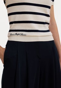 Kremna in mornarsko modra črtasta pletena majica s kratkimi rokavi, rebrastim robom in izvezenim logotipom "Lauren Ralph Lauren" na pasu, v kombinaciji z mornarsko modrimi hlačami.