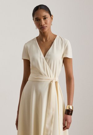 Femeie purtând o rochie crem cu mâneci scurte, în stil petrecut, cu un nod lateral, cercei mari aurii și două brățări la încheietura mâinii drepte.