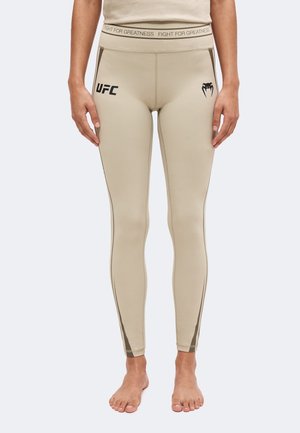 Leggings beige UFC avec texte "Fight for Greatness" à la taille et un logo serpent noir sur la cuisse droite, portés par une personne debout pieds nus.