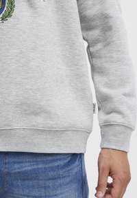 Sudadera gris con puños y dobladillo de canalé, que presenta un parche con logo en el lateral. El material parece suave y texturizado. Se lleva con jeans de mezclilla azul.