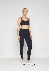 Schwarzer Sport-BH mit verstellbaren Trägern, kombiniert mit hoch taillierten schwarzen Leggings. Weiße Sportschuhe und eine schwarze Wasserflasche sind ebenfalls enthalten.