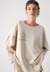 Beige oversized sweatshirt met lange mouwen, met grote crèmekleurige tekst op de voorkant. Gladde textuur en een relaxte pasvorm.