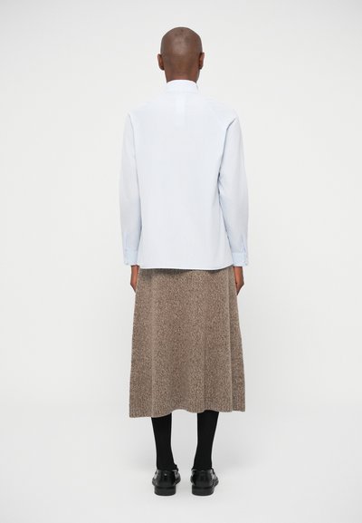 Svetlo modra srajca z dolgimi rokavi in sproščenim krojem, v kombinaciji z rjavo teksturirano krilo do kolen in črnimi čevlji, gledano zadaj.