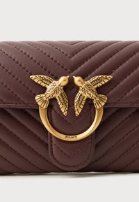Borsa a tracolla in pelle trapuntata color bordeaux con un'applicazione di un uccello in tono oro e hardware circolare, caratterizzata da evidenti dettagli di cucitura.