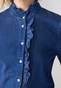 Denimskjorta i mörkblått, med en flärd på framsidan och en hög krage. Vita knappar är jämnt fördelade längs öppningen.