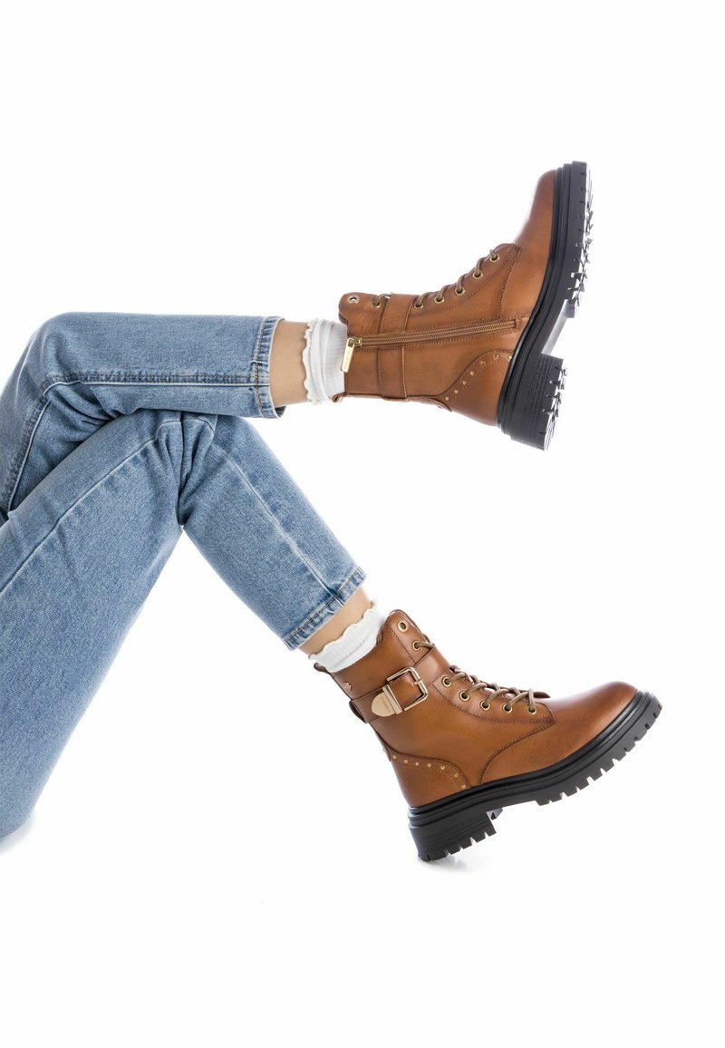 Botas de tobillo de cuero marrón con cordones en la parte delantera, detalle de hebilla y acentos de tachuelas. Suela negra gruesa que proporciona tracción. Llevadas con jeans de color azul claro.