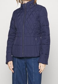 Granatowa pikowana kurtka z wysokim kołnierzem, zapinana na zamek z przodu, z dwoma bocznymi kieszeniami. Charakteryzuje się teksturowaną tkaniną i przeszywanym wzorem w romby.